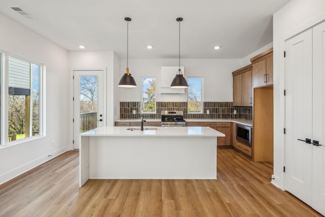 kitchen featuring appliances with stainless steel finishes, a kitchen island with sink, sink, pendant lighting, and light hardwood / wood-style flooring