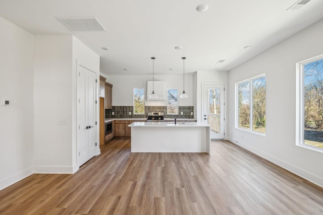 kitchen with backsplash, a kitchen island with sink, light hardwood / wood-style flooring, stainless steel range, and decorative light fixtures