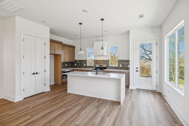 kitchen with backsplash, a wealth of natural light, custom exhaust hood, hanging light fixtures, and an island with sink