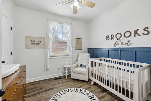 bedroom with a crib, dark wood-type flooring, and ceiling fan