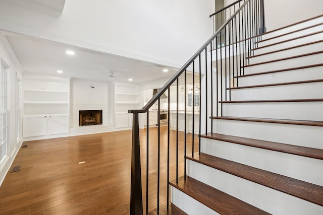 stairway featuring ceiling fan, built in features, crown molding, wood-type flooring, and a fireplace