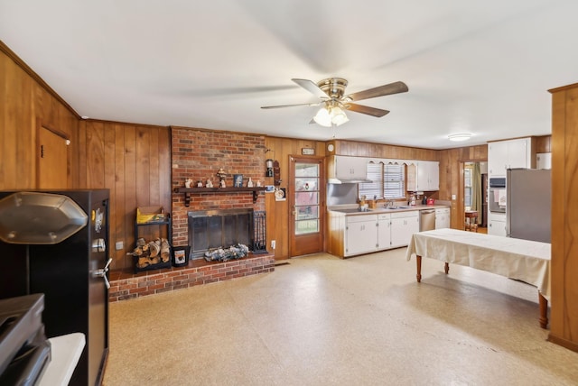 kitchen featuring white cabinets, wooden walls, a brick fireplace, ceiling fan, and stainless steel appliances