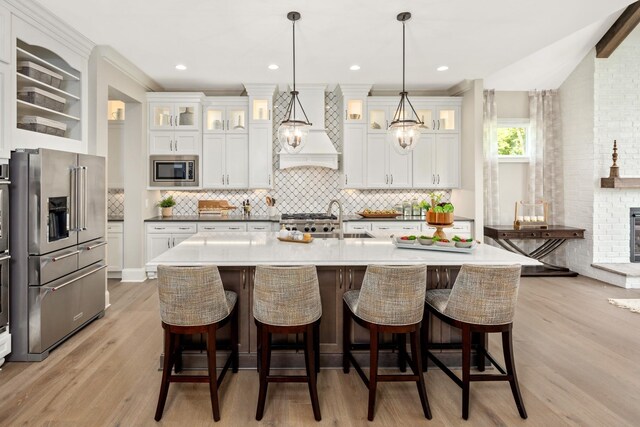 kitchen featuring custom exhaust hood, light wood-type flooring, appliances with stainless steel finishes, decorative light fixtures, and white cabinetry