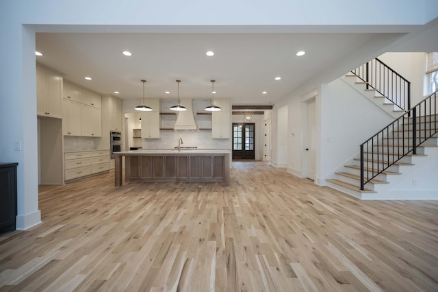 kitchen with double oven, a kitchen island with sink, sink, decorative light fixtures, and light hardwood / wood-style flooring