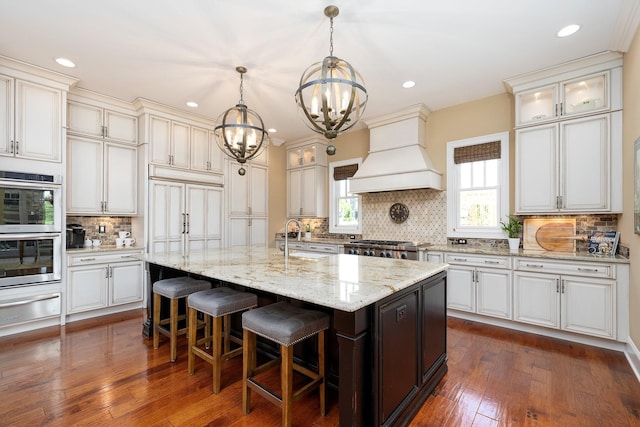 kitchen featuring tasteful backsplash, custom exhaust hood, stainless steel double oven, a kitchen island with sink, and pendant lighting