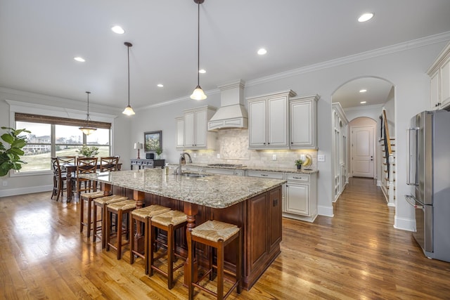 kitchen featuring premium range hood, hanging light fixtures, stainless steel appliances, light stone countertops, and an island with sink