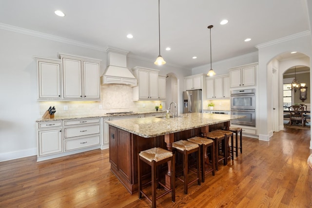 kitchen with premium range hood, white cabinetry, sink, stainless steel appliances, and a center island with sink