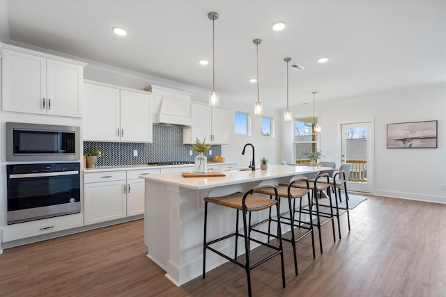 kitchen with appliances with stainless steel finishes, custom range hood, a center island with sink, white cabinets, and hanging light fixtures