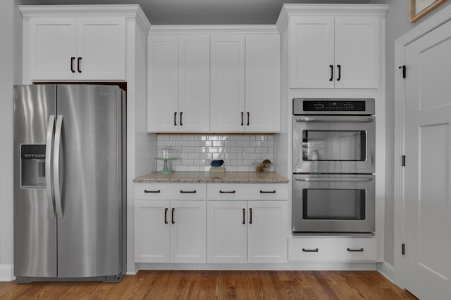 kitchen with appliances with stainless steel finishes, light wood-type flooring, backsplash, light stone counters, and white cabinetry