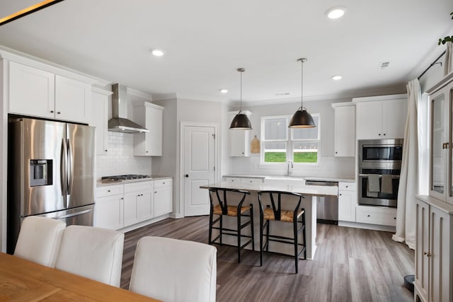 kitchen featuring a center island, wall chimney exhaust hood, hanging light fixtures, stainless steel appliances, and white cabinets