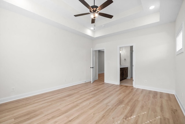 unfurnished bedroom featuring a tray ceiling, ensuite bath, ceiling fan, and light wood-type flooring