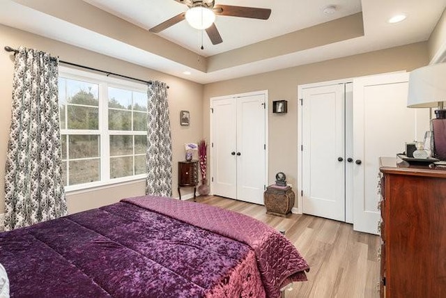 bedroom with ceiling fan, a tray ceiling, light wood-type flooring, and two closets