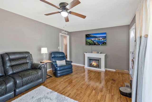 living area featuring a ceiling fan, a glass covered fireplace, baseboards, and wood finished floors