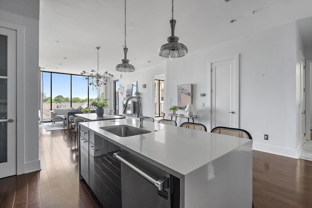 kitchen featuring stainless steel dishwasher, expansive windows, a kitchen island with sink, sink, and hanging light fixtures