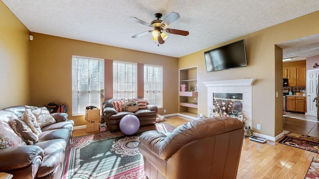 living room featuring a tile fireplace, ceiling fan, light hardwood / wood-style floors, a textured ceiling, and built in shelves