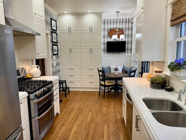 kitchen with stainless steel appliances, sink, wall chimney range hood, decorative light fixtures, and white cabinetry