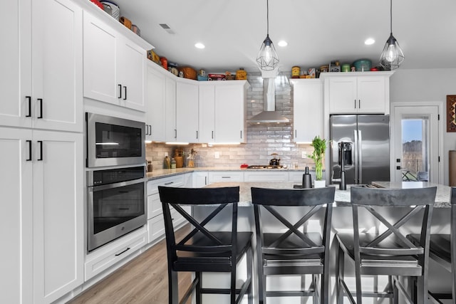 kitchen with wall chimney exhaust hood, white cabinetry, stainless steel appliances, and decorative light fixtures