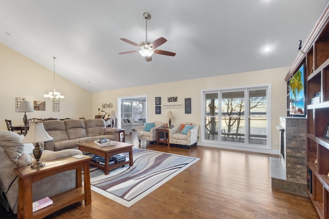 living room with lofted ceiling, a stone fireplace, wood-type flooring, and ceiling fan with notable chandelier