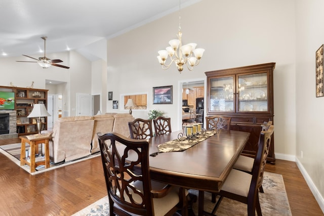 dining space featuring ceiling fan with notable chandelier, a stone fireplace, wood-type flooring, and high vaulted ceiling