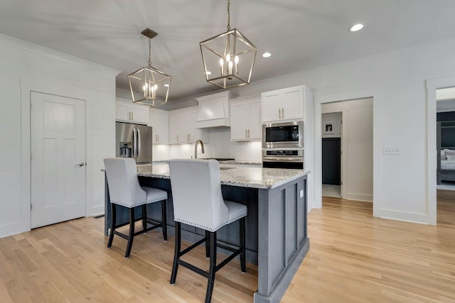 kitchen featuring custom exhaust hood, a kitchen island with sink, appliances with stainless steel finishes, decorative light fixtures, and white cabinetry