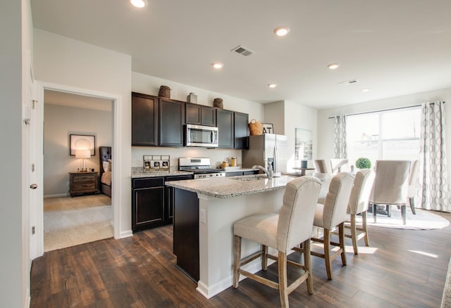 kitchen with appliances with stainless steel finishes, light stone counters, a breakfast bar, a kitchen island with sink, and dark wood-type flooring