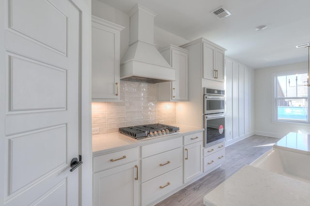 kitchen featuring stainless steel appliances, light hardwood / wood-style floors, decorative backsplash, white cabinets, and custom range hood