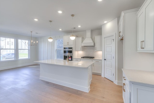 kitchen with double oven, an island with sink, decorative light fixtures, white cabinets, and custom exhaust hood