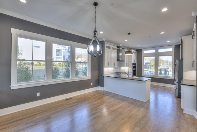 kitchen featuring kitchen peninsula, stainless steel appliances, wall chimney range hood, decorative light fixtures, and white cabinetry