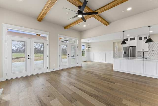 unfurnished living room with beam ceiling, french doors, a healthy amount of sunlight, ceiling fan with notable chandelier, and hardwood / wood-style flooring