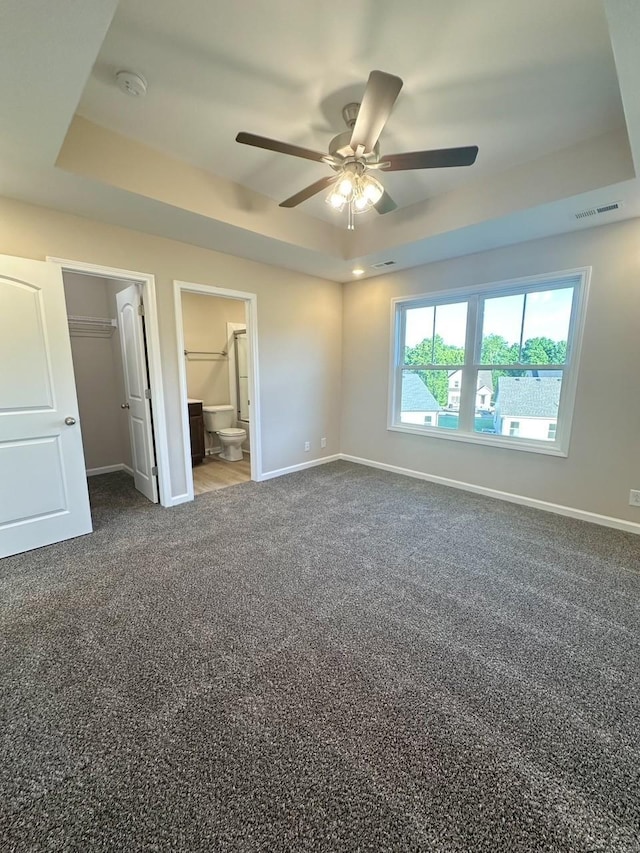 unfurnished bedroom featuring a tray ceiling, connected bathroom, ceiling fan, and dark carpet