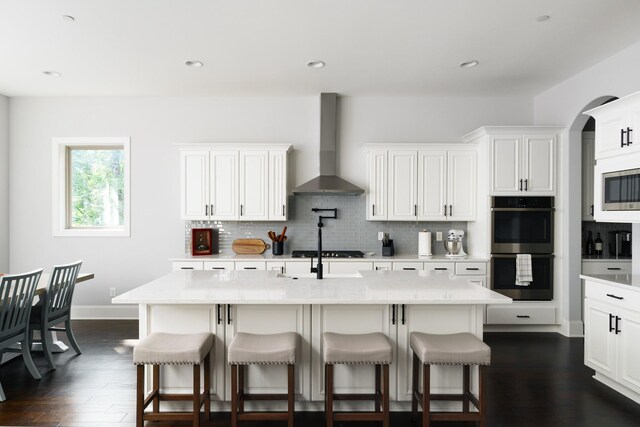 kitchen featuring a kitchen island with sink, white cabinets, wall chimney range hood, tasteful backsplash, and stainless steel appliances