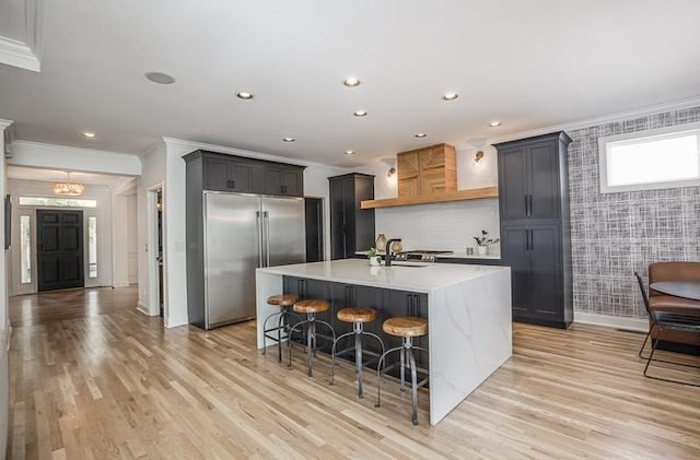 kitchen with a kitchen bar, ornamental molding, stainless steel built in refrigerator, light hardwood / wood-style floors, and an island with sink