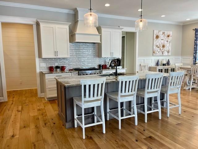 kitchen featuring a kitchen island with sink, white cabinets, decorative light fixtures, and custom range hood