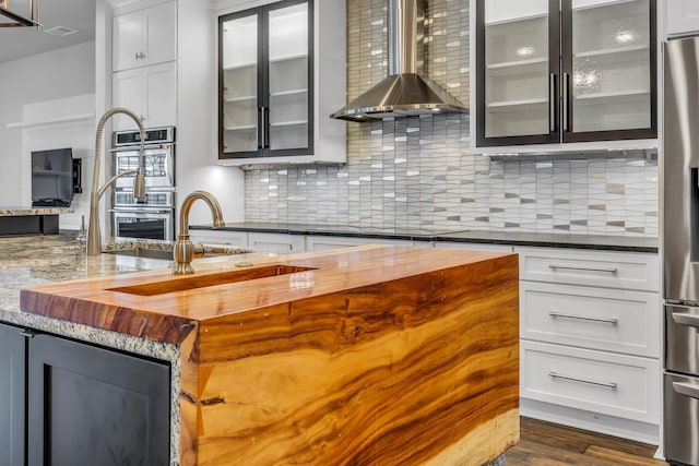 kitchen with butcher block counters, wall chimney range hood, appliances with stainless steel finishes, tasteful backsplash, and white cabinetry