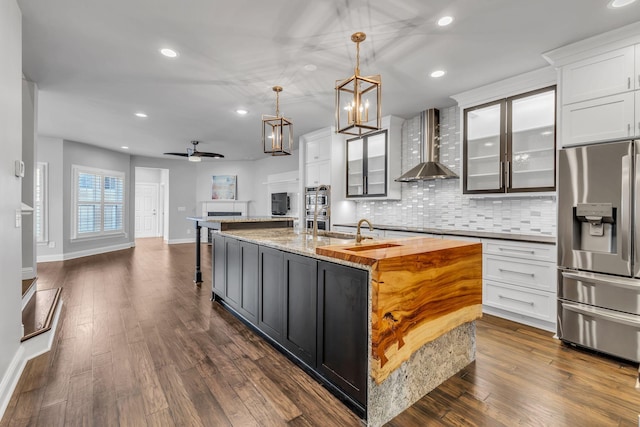 kitchen with appliances with stainless steel finishes, ceiling fan, wall chimney range hood, pendant lighting, and a center island with sink