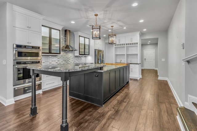 kitchen featuring light stone counters, wall chimney exhaust hood, stainless steel appliances, a spacious island, and white cabinets