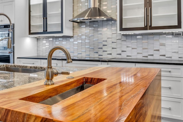 kitchen featuring decorative backsplash, wall chimney exhaust hood, sink, butcher block countertops, and white cabinetry