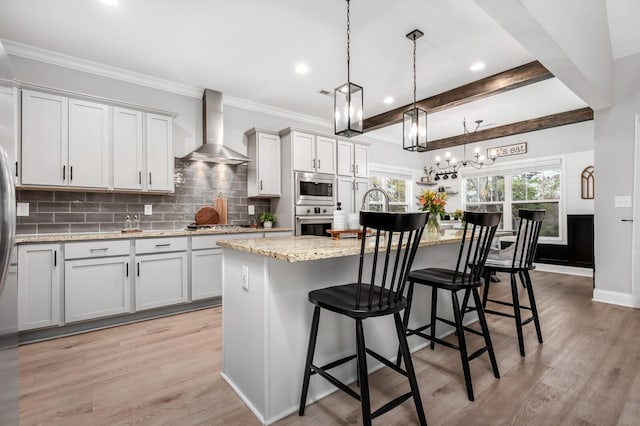 kitchen with appliances with stainless steel finishes, a breakfast bar, a kitchen island with sink, wall chimney range hood, and white cabinetry