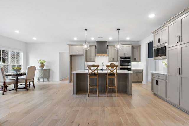 kitchen featuring built in microwave, an island with sink, gray cabinetry, hanging light fixtures, and wall chimney range hood