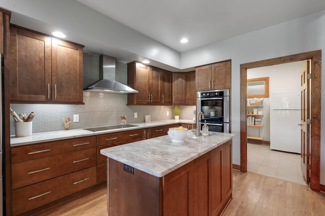 kitchen featuring wall chimney range hood, black electric cooktop, an island with sink, double oven, and white fridge