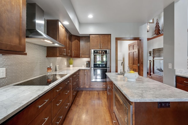 kitchen with stainless steel appliances, wall chimney range hood, tasteful backsplash, a kitchen island with sink, and light wood-type flooring