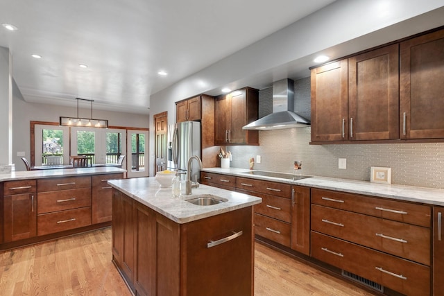 kitchen with stainless steel fridge, black electric stovetop, sink, wall chimney range hood, and an island with sink