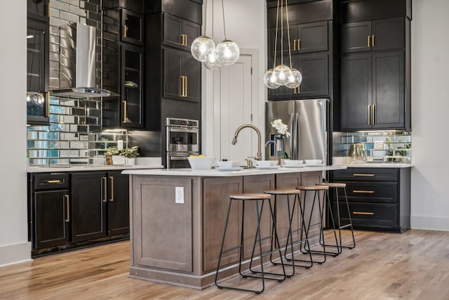 kitchen with stainless steel appliances, ventilation hood, an island with sink, pendant lighting, and light wood-type flooring