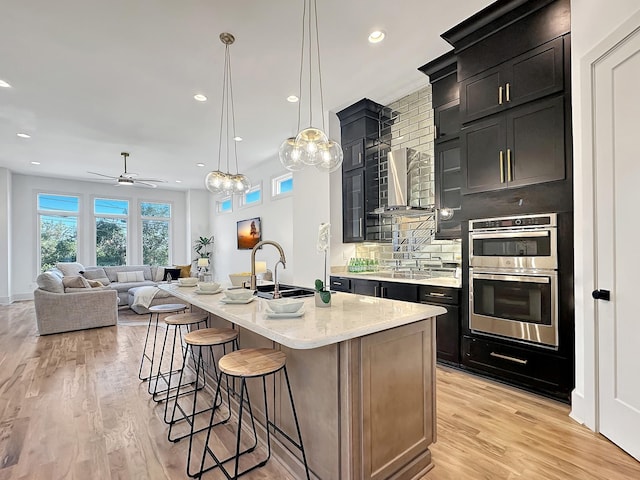 kitchen with sink, hanging light fixtures, wall chimney range hood, double oven, and an island with sink