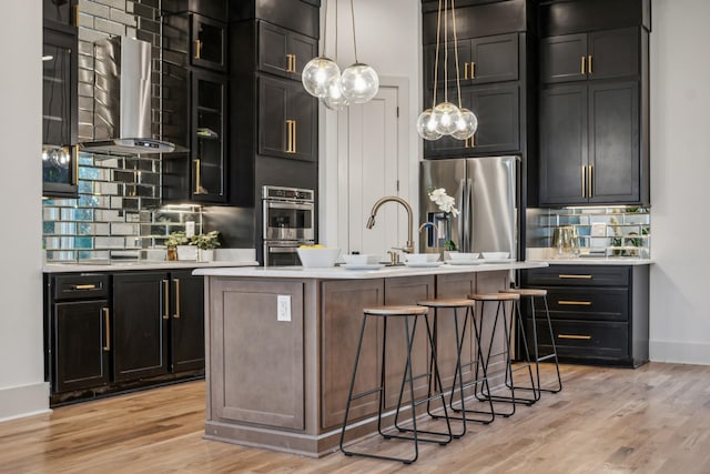 kitchen featuring appliances with stainless steel finishes, ventilation hood, decorative light fixtures, light hardwood / wood-style flooring, and an island with sink