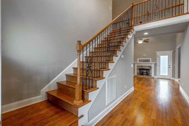 stairway featuring ceiling fan, a stone fireplace, wood-type flooring, and a high ceiling