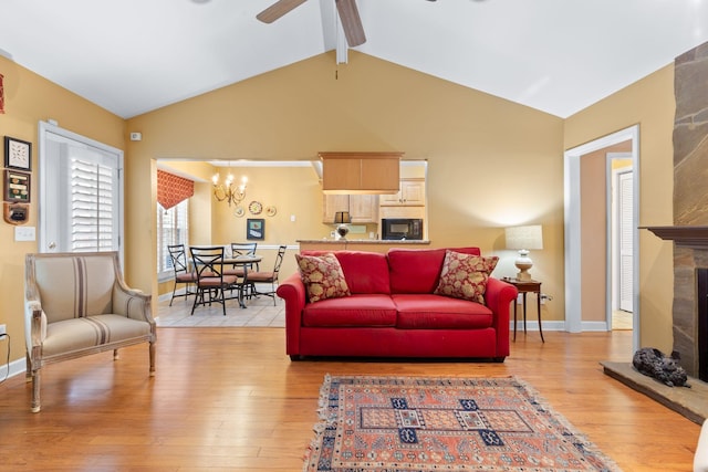 living room with lofted ceiling with beams, a stone fireplace, light wood-type flooring, and ceiling fan with notable chandelier