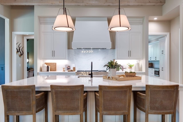 kitchen featuring pendant lighting, ventilation hood, white cabinets, a center island with sink, and decorative backsplash