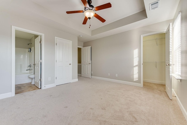 unfurnished bedroom with a raised ceiling, connected bathroom, ceiling fan, and light colored carpet