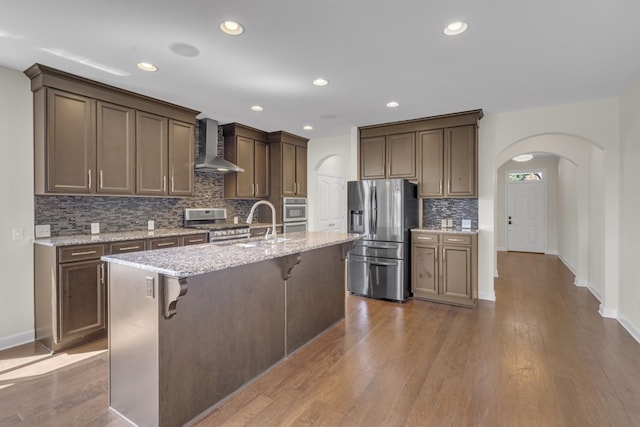 kitchen with a kitchen bar, stainless steel appliances, wall chimney range hood, wood-type flooring, and a center island with sink